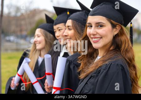 Group of multi ethnic female graduates wearing academic gowns and holding diplomas, smiling outdoors Stock Photo