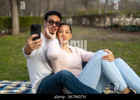 Young couple making playful and silly faces while capturing a selfie with their smartphone, enjoying a joyful day in the park Stock Photo