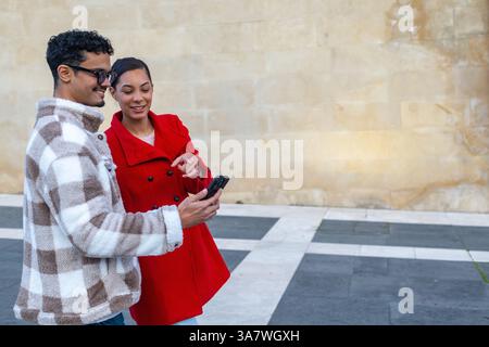 Young man and woman smiling while using a smartphone, enjoying a vibrant day together in the city, embracing urban lifestyle and connection Stock Photo