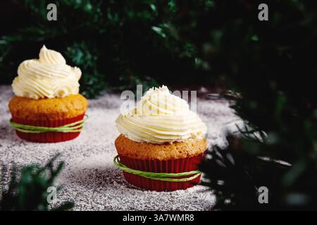 Yummy colorful cupcakes with whipped cream in red paper cups decorated with green rope placed near Christmas tree branches on table sprinkled with sug Stock Photo