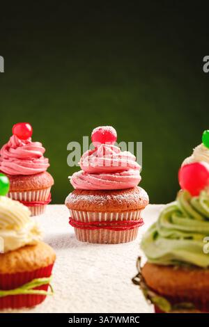 Yummy festive Christmas cupcakes decorated with colorful frosting and cherry berries placed on table sprinkled with white sugar powder against green b Stock Photo