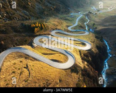 Aerial view of winding mountain road, river in alpine valley at sunset in autumn. Top view of road in swiss mountain pass, yellow grass, trees and sun Stock Photo
