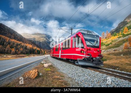Red modern passenger train is moving on railway in swiss alps at sunset in autumn.  Landscape with train in high mountains, road, orange trees in fall Stock Photo