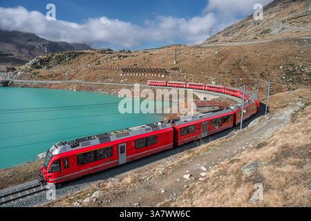 Aerial view of modern red train near azure mountain lake and dramatic cloudy sky at sunset in autumn. Bernina Express, Switzerland. Top view of train Stock Photo