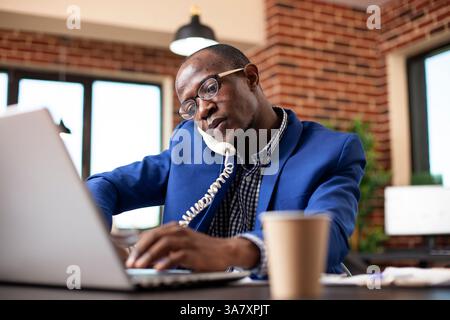 Financial advisor discussing updates with client on landline phone and working on laptop at office desk. Closeup of black businessman handling calls and typing on device at brick wall workspace. Stock Photo