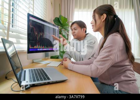 A young man and woman sit at a wooden desk, both focused on a large computer screen displaying detailed market graphs. Stock Photo
