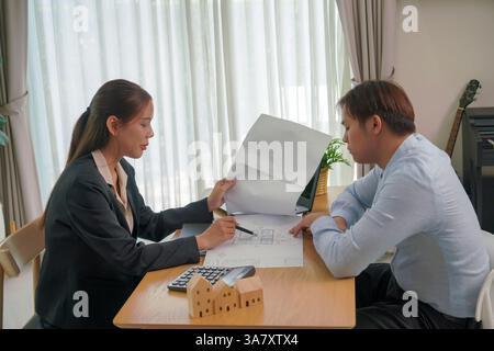 businesswoman in a suit sits across from a man, presenting architectural plans on paper. With a small wooden house model and a calculator on the table Stock Photo