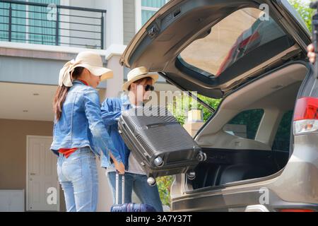 young man in a denim jacket lifts a large suitcase into the trunk of a car while a woman, also dressed in denim and a wide-brimmed hat, stands nearby Stock Photo