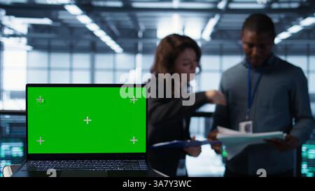 Green screen laptop used by data center admins in blurry background reviewing equipment diagnostics. Chroma key notebook used by coworkers adjusting server room configurations, camera B Stock Photo