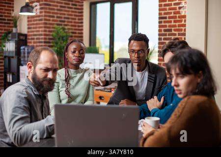 Multiethnic team gathers around laptop for discussion, showcasing focus and teamwork in modern workspace. Startup employees collaborating on project with concentration at shared device on office desk. Stock Photo