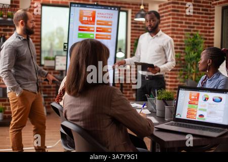 Bearded businessmen stand near digital screen, presenting marketing strategies at group meeting. Black male entrepreneur with clipboard leads business project presentation at brick wall office. Stock Photo