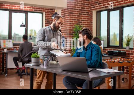 Caucasian man sits with laptop as male coworker checks clipboard and explains updates on market analysis. Bearded businessmen are actively collaborating at office desk, planning project presentation. Stock Photo