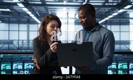 Engineers using laptop in data center, managing server virtualization, running scripts. IT specialists using programming language on notebook in server hub, finetuning network protocols, camera B Stock Photo