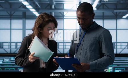 Data center management executives reading paperwork files, reviewing system diagnostics. Server farm colleagues looking over maintenance schedules documents and cybersecurity protocols, camera A Stock Photo