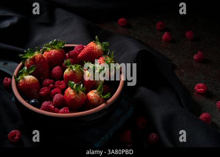 Black ceramic pot filled with red berries, strawberries, raspberries, and blueberries on a dark wooden board on a black table with a black tablecloth Stock Photo