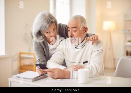 Senior woman hugging husband sitting at desk and showing video on mobile phone at home. Stock Photo