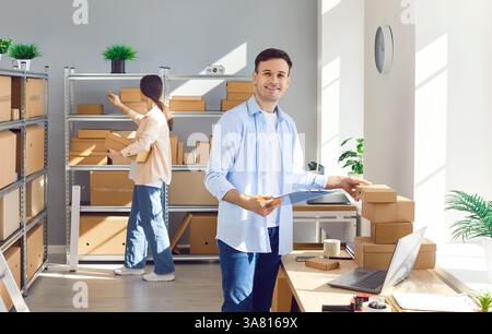 Man standing in warehouse, doing inventory, looking at camera, woman inspecting boxes and parcels. Stock Photo
