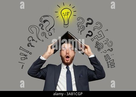 Emotional shocked businessman in suit with laptop on his head on grey background and drawing of light bulb and question marks Stock Photo