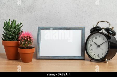 Stylish home decor with an empty frame, alarm clock, and potted plants on a wooden surface against a textured wall Stock Photo