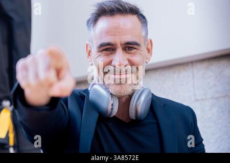 Smiling businessman with headphones pointing at camera while standing outdoors in style Stock Photo