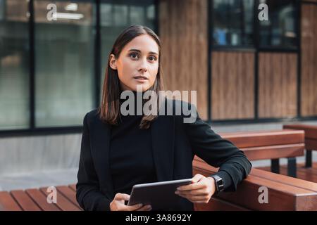 Portrait of young businesswoman in black stylish attire sitting outdoors with a digital tablet Stock Photo