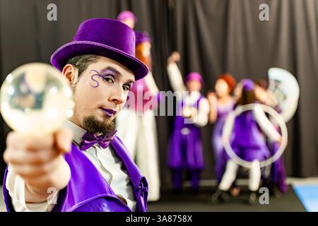 Magician wearing purple top hat and suit showing crystal ball with circus performers juggling and dancing in background Stock Photo