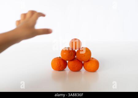 A child's hand reaches for a pyramid of vibrant tangerines on a pristine white surface. The juicy citrus fruits are playfully stacked, tempting a grab Stock Photo