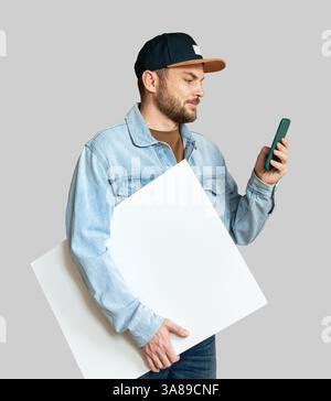 Man in denim jacket and snapback holding blank white board while using smartphone Stock Photo