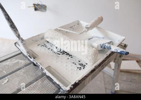 A paint tray with white paint, roller, and brush sits on a ladder platform, ready for painting walls during a room renovation. Stock Photo