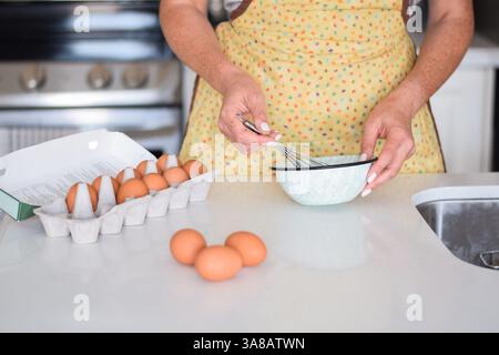 Woman wearing yellow apron is whisking eggs in a bowl on kitchen counter at home Stock Photo