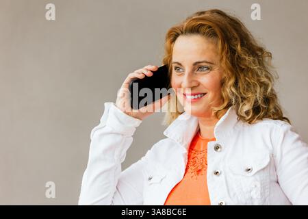 cheerful middle-aged woman in orange top and white jacket holding a smartphone to her ear, smiling while having a pleasant conversation against a plai Stock Photo
