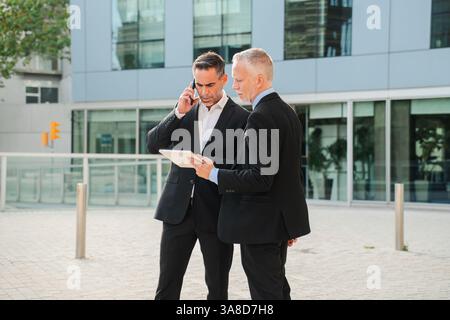 Two businessmen engaged in a serious conversation as one executive talks on the phone while the other examines details on a tablet device, showcasing professionalism and collaboration in business. High quality photo Stock Photo