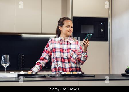 Young woman in casual plaid shirt standing in kitchen room using her mobile phone. Checking status of meal delivery or reading dinner recipe Stock Photo