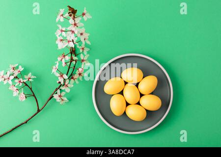 Yellow Easter eggs on grey plate with cherry blossom branch on green background. Top view, flat lay. Stock Photo