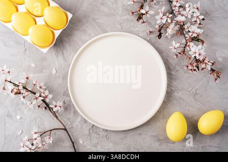 Yellow Easter eggs on gray background with empty plate and cherry blossom branches. Top view, flat lay, mockup. Stock Photo