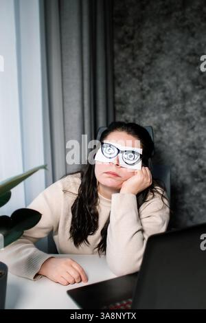 Tired Caucasian woman freelancer feeling stressed from work problems. Irritation burnout. Painted eyes on adhesive notes glued to face. Not having eno Stock Photo