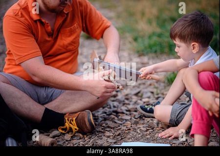 A father guides his young son in using a saw to cut a small branch. The outdoor setting suggests a hands-on learning experience, camping, or bushcraft Stock Photo