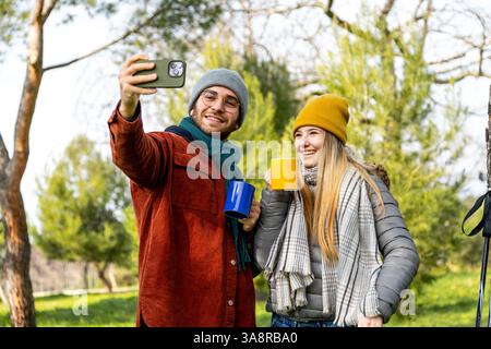 Young couple taking selfie with smartphone while enjoying hot drinks during hike in a forest Stock Photo