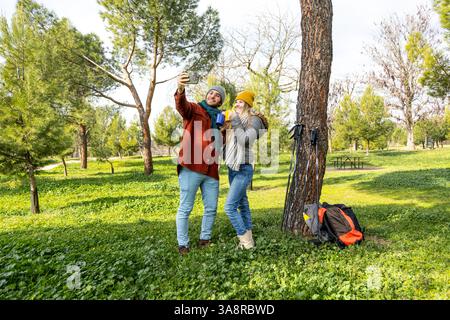 Hikers taking a selfie with mobile phone in a park while drinking hot beverages during a break Stock Photo
