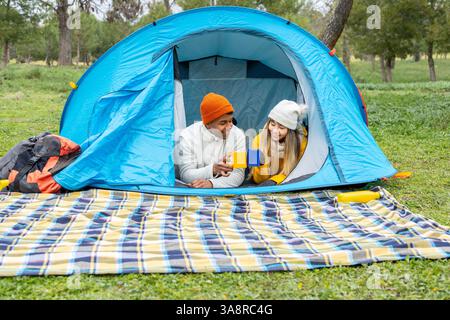 Interracial couple lying inside their tent, toasting with mugs and enjoying a hot drink on a camping trip Stock Photo