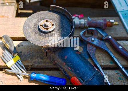 A close-up of a used angle grinder with a worn disc, alongside various hand tools like pliers and screwdrivers, all resting on a wooden surface, sugge Stock Photo