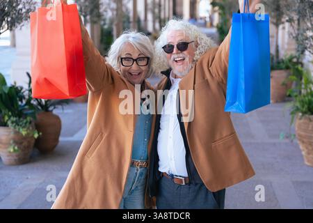 Happy senior couple holding shopping bags and celebrating outdoors in city street Stock Photo