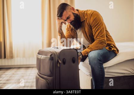 Man in his 30s sitting on bed in hotel room. He is tired and exhausted. Suitcase is beside the bed. Stock Photo