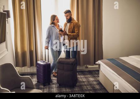 Couple in their 30s standing in hotel room with their suitcases unpacked. They are using smartphone and planning their stay. Stock Photo