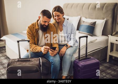 Happy married couple sitting on bed in hotel room and planning their vacation by using smartphone. Suitcases are beside the bed. Stock Photo