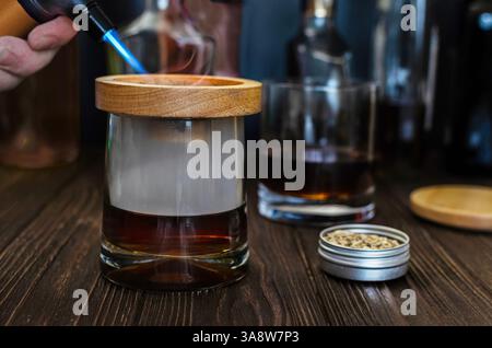 A close-up shot of a cocktail being smoked using a handheld torch and wood chips. The smoke is captured inside a glass with a wooden topper, showcasin Stock Photo