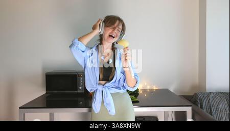 Young woman singing in kitchen, using potato as microphone, wearing Stock Photo