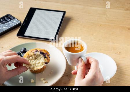A person is eating a muffin and holding a cup of coffee. The scene is set on a wooden table with a reading device and mobile phone Stock Photo