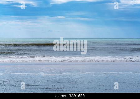 Gentle waves roll toward the shore under a cloudy blue sky. Stock Photo