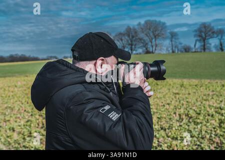 A man wearing a black jacket and hat is taking a picture of a field. He is holding a camera and he is focused on capturing the scene. The field is gre Stock Photo
